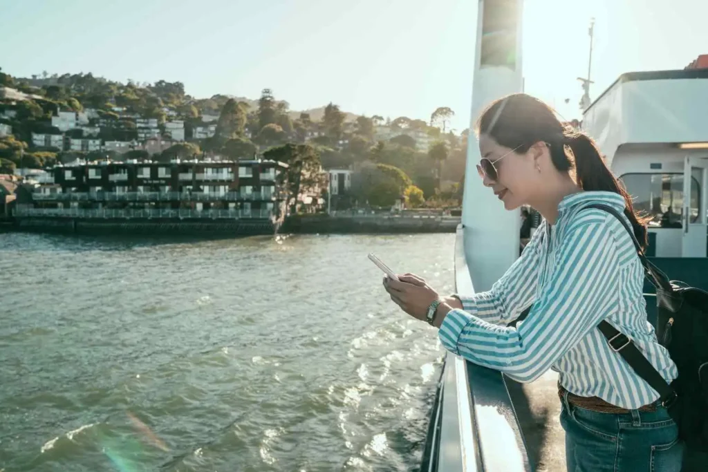A female tourist on a boat ferry using her phone to learn how to book a private ride from Cruise to Disney with coastal buildings in the background.