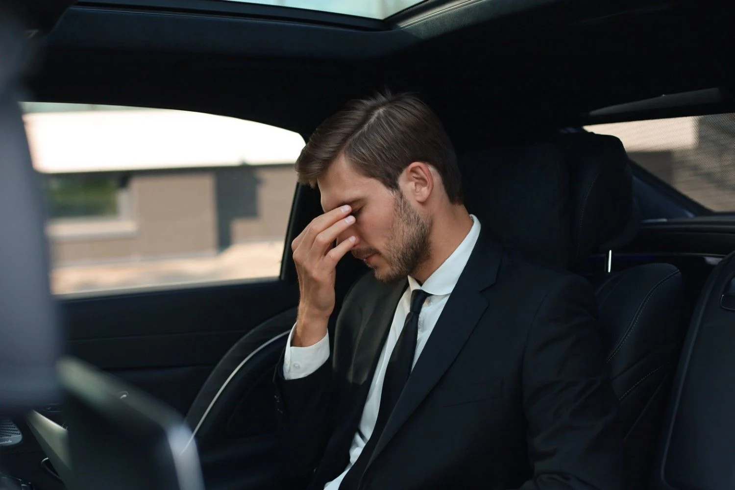An exhausted businessman in a suit sitting in the backseat of a car and pinching the bridge of his nose, highlighting the importance of understanding travel fatigue during long business trips.